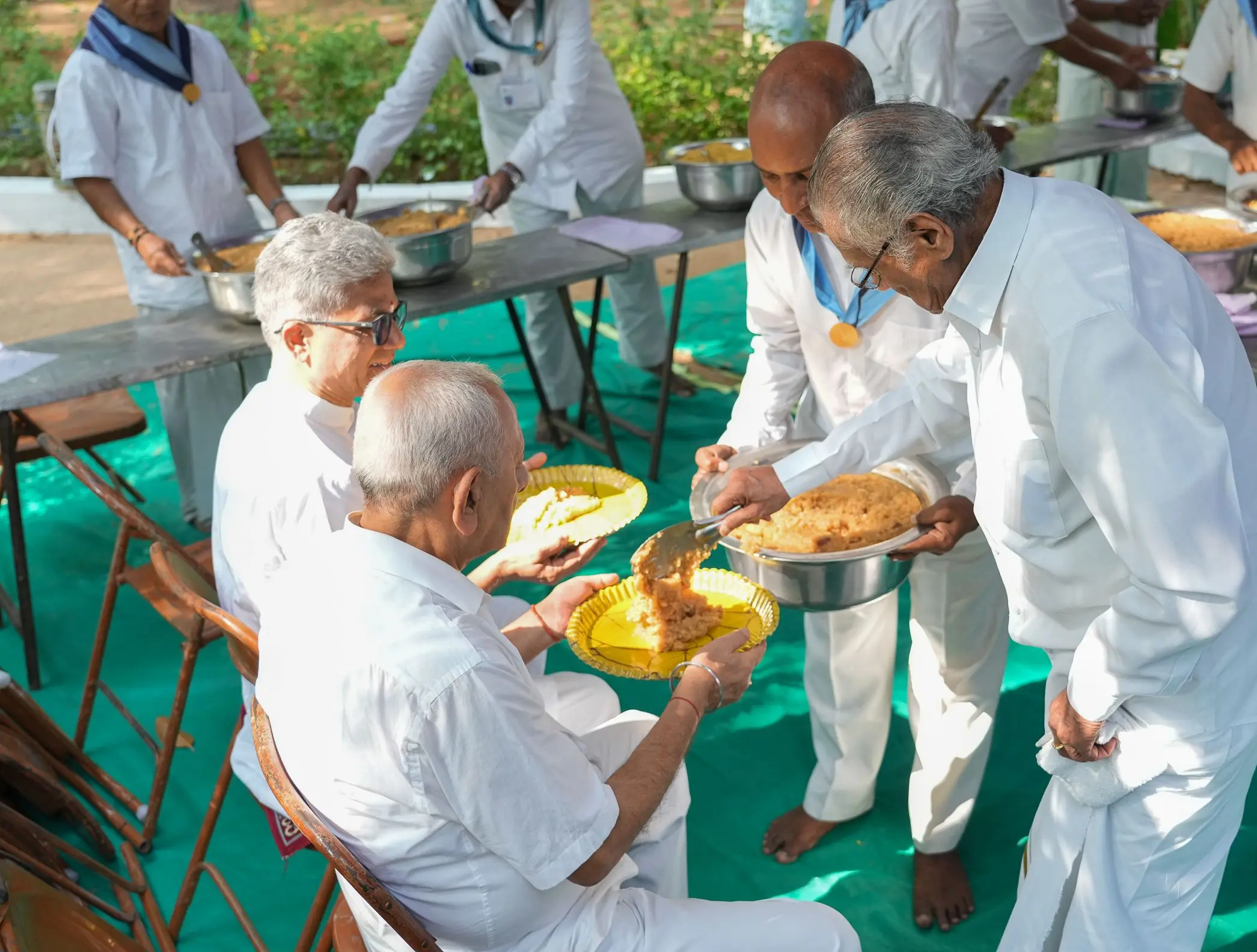 मेज़ों पर बैठे बुज़ुर्ग अतिथियों को प्रसाद परोसते सेवादल स्वयंसेवक