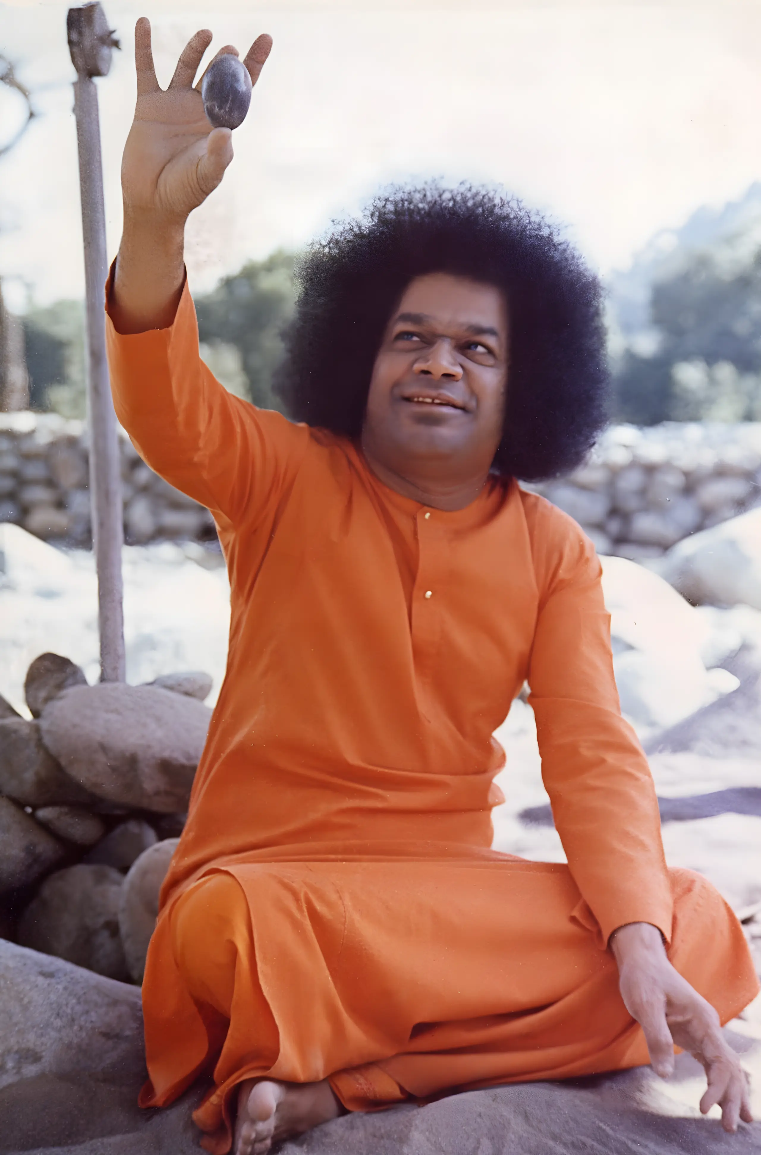 नदी किनारे चट्टानों पर बैठे, आशीर्वाद में हाथ उठाए भगवान श्री सत्य साई बाबा
